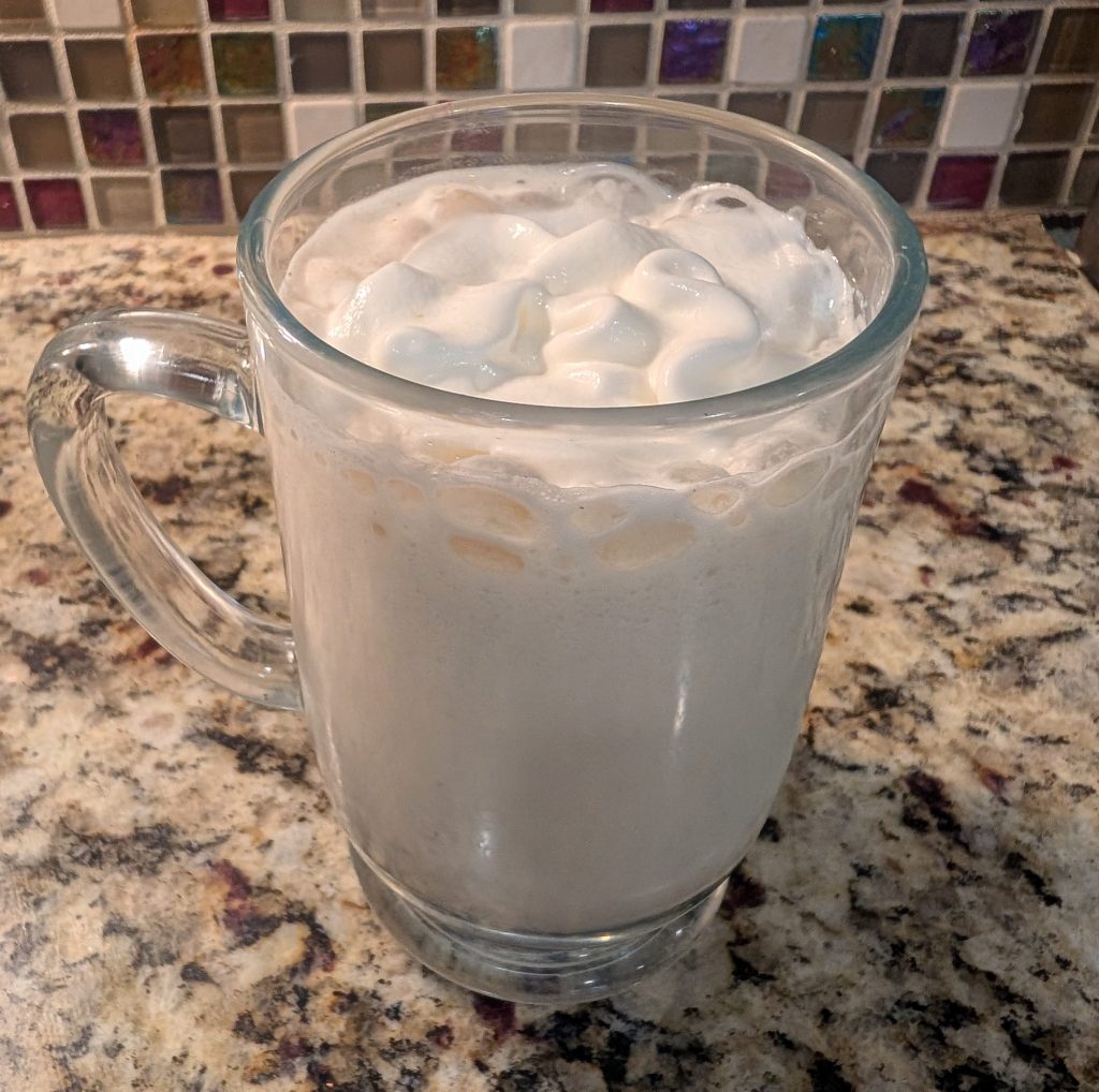 A clear glass mug filled with a creamy beverage topped with whipped cream, placed on a textured countertop.
