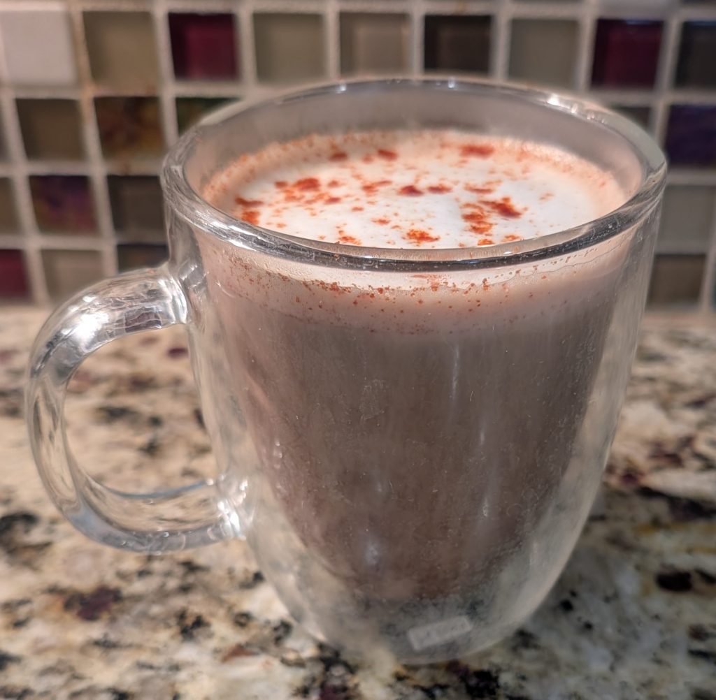 A clear glass mug filled with frothy hot chocolate, topped with a sprinkle of cinnamon, set on a granite countertop.